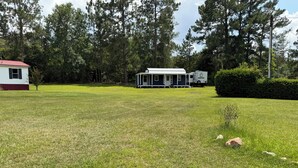 Une zone herbeuse avec un bâtiment blanc et une maison mobile en arrière-plan, le tout entouré d’arbres.
