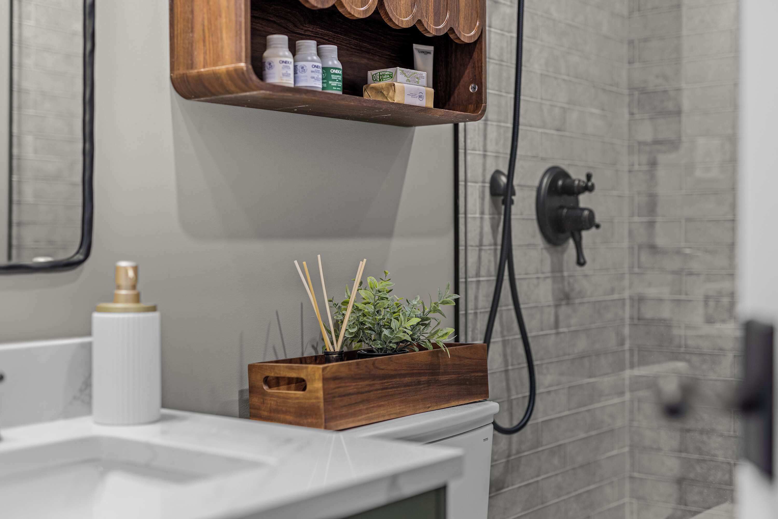 Une salle de bain avec une étagère en bois où sont rangés les articles de toilette, un lavabo blanc et une douche avec un tuyau noir.