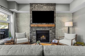 A living room with a stone fireplace, two armchairs, a sofa, a lamp, and a view of the outdoors. - BRAND NEW Luxury Downtown Condo Settlers on Main (Canmore)