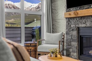 Een gezellige woonkamer met een open haard, een comfortabele fauteuil, een klein bijzettafeltje met een decoratie, en uitzicht op bergen door grote ramen.