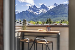 Un balcon donnant sur des montagnes enneigées, une table avec des chaises et une fenêtre offrant une vue sur une petite ville.