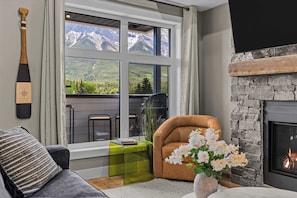 Un salon chaleureux avec une cheminée, une grande fenêtre donnant sur les montagnes, un fauteuil en cuir et une petite table d’appoint verte.