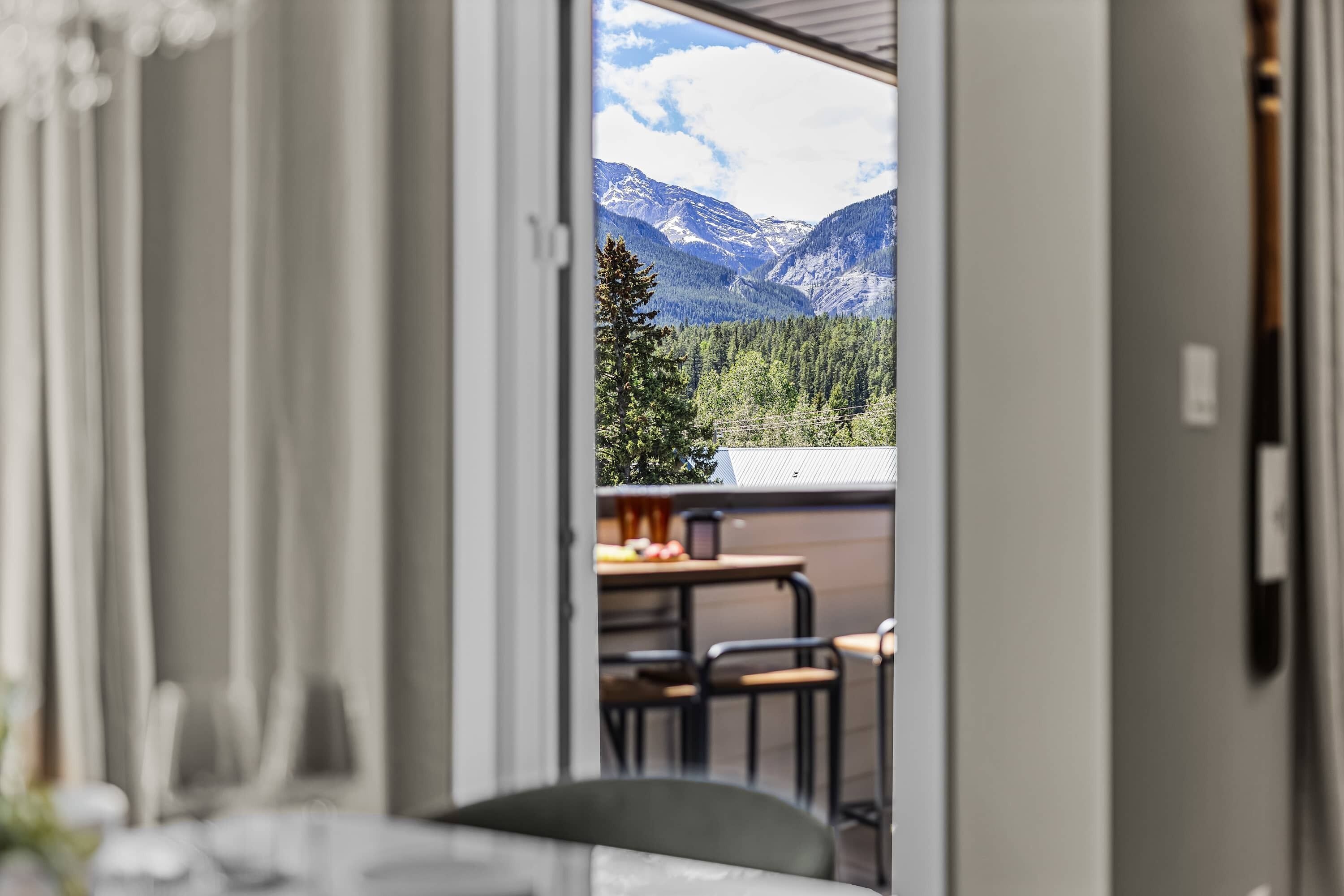Une vue intérieure d’une pièce avec une table et des chaises, une fenêtre donnant sur un paysage montagneux et une porte menant à l’extérieur.