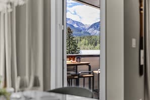 Une vue intérieure d’une pièce avec une table et des chaises, une fenêtre donnant sur un paysage montagneux et une porte menant à l’extérieur.