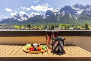 Une table en bois sur laquelle est disposée une assiette de fruits, deux verres remplis de boisson et une lanterne, le tout sur une terrasse offrant une vue sur la montagne.