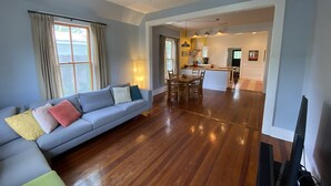 A living room with a sofa, wooden flooring, and a kitchen in the background. - Gorgeous Zilker Cottage - Monthly Rental - Available August (Austin)