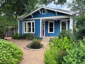 Ein blaues Haus mit einer Veranda, ein kleiner Garten mit verschiedenen Pflanzen und ein Kiesweg, der zur Tür führt.