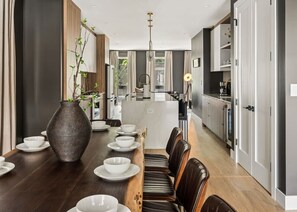 A modern dining area with a long wooden table set for a meal, flanked by dark leather chairs, and a large vase of greenery.