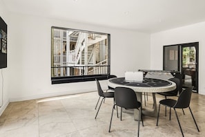 A modern room with a round table, black chairs, a foosball table, and a large window with a view of a white building and staircase. - 2,400 SF 4BR + 2 Parking | Steps to Wrigley (Chicago)