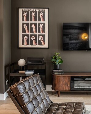 A living room with a leather armchair, a framed baseball pitches poster, a television, and a bookshelf.