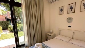 Hotellihuone, jossa on sänky, yöpöytä, ilmastointilaite ja liukuovet, jotka johtavat ulkotilaan.