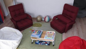Una habitación con dos sillas rojas, un sillón de mimbre blanco, una mesita con libros y una alfombra colorida.