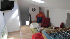Una habitación con mesa de fútbol, un sillón de peluche rojo, una silla roja, televisor montado en la pared y dardo.