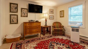 A living room with a flat-screen TV, a wooden dresser, a patterned armchair, a standing lamp, and a large window with blinds.