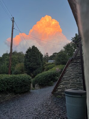 A stone pathway leading to a building with a stone roof, surrounded by greenery and a large cloud in the sky.