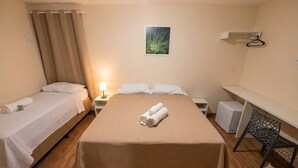 Habitación de hotel con cama, dos mesitas de noche, lámpara, un mueble blanco pequeño, una silla y una estantería con perchero.