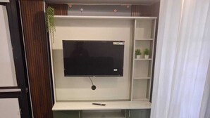 Une pièce avec un téléviseur fixé au mur au-dessus d’un meuble blanc, un canapé bleu et une petite étagère avec des plantes.