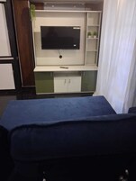 Una habitación con un televisor montado en un mueble blanco, un sofá azul y un estante pequeño con plantas.