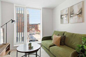 Un salon moderne avec un canapé vert, une table basse ronde, un lampadaire et une grande fenêtre équipée de stores.