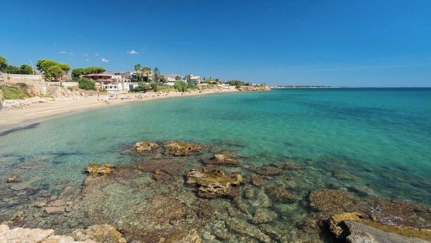 A coastal beach with clear water, rocky shoreline, and buildings in the background. - La Dimora di Vanna by Rentbeat (Avola)