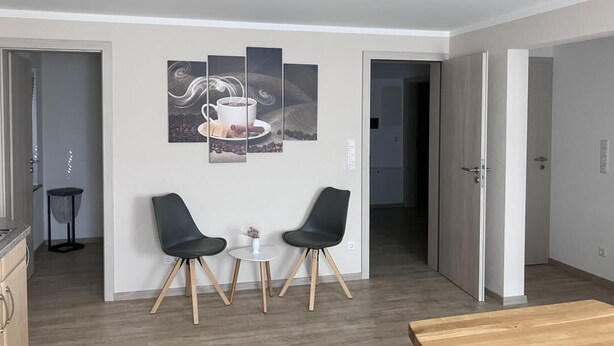 A modern living room with a dining area, featuring a coffee-themed artwork on the wall, two black chairs, a small round table, and a wooden dining table. - NEW! To the Old Inn (Reich)