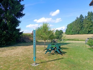 Eine begrünte Außenfläche mit einer Metallkonstruktion, einem kleinen Baum und einem Metalltisch mit Stühlen.
