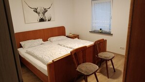 A bedroom with a wooden bed, two chairs, a nightstand, a wall-mounted shelf with a plant, and a window with blinds. - NEW! To the Old Inn (Reich)