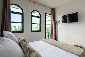 Een slaapkamer met een bed, twee ramen met gordijnen, een televisie aan de muur en uitzicht op gebouwen.