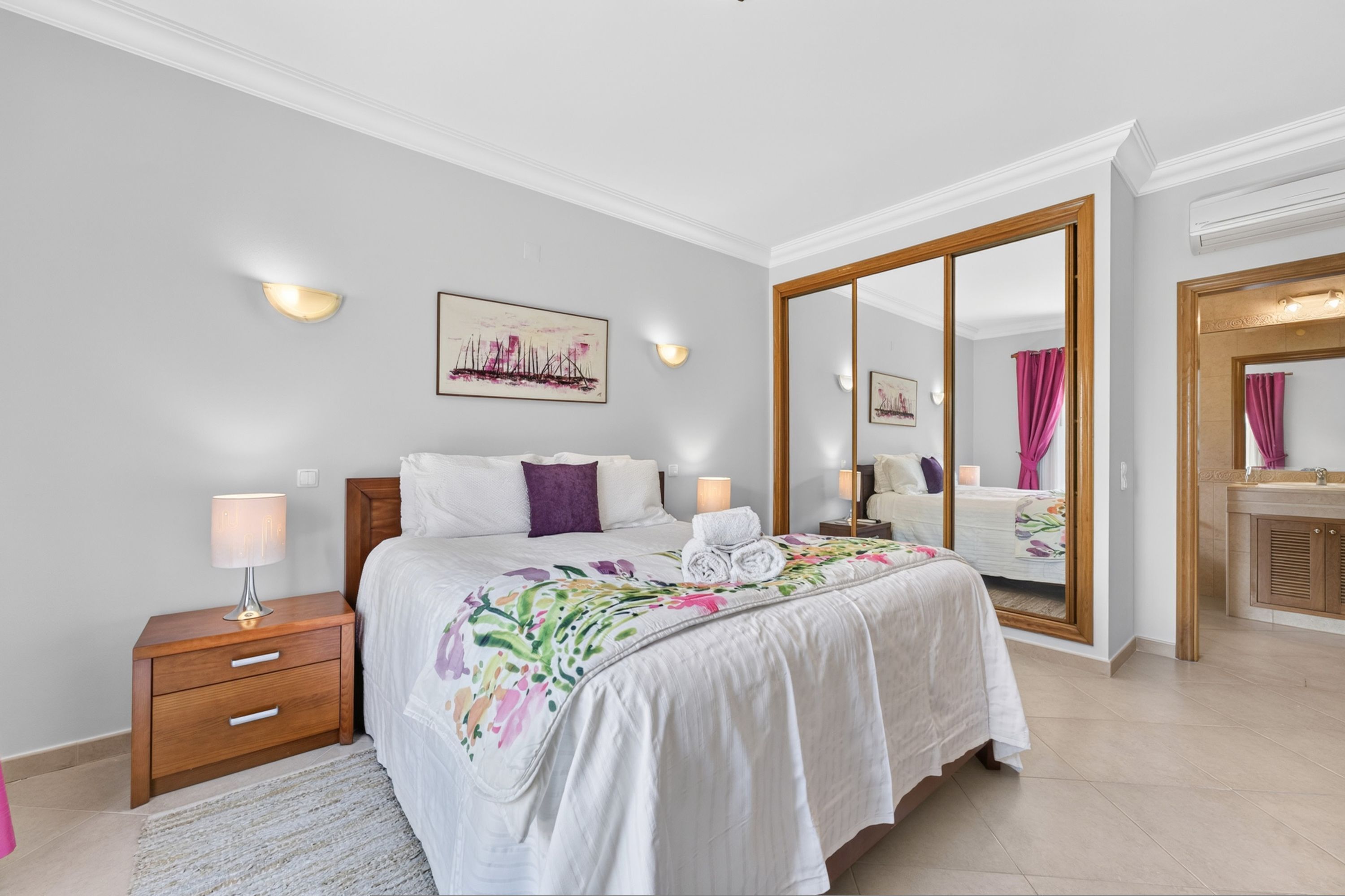 Un dormitorio con cama, mesitas de noche, un espejo, un cuadro y un baño con lavamanos y espejo.