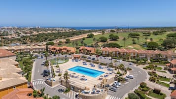 Zona residencial con piscina rodeada de sillas de descanso y sombrillas, una calle con coches y vista al océano.