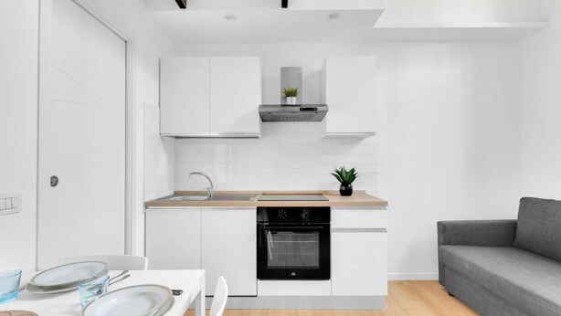 Una cocina moderna con armarios blancos, horno empotrado y un sofá gris en la sala de estar.