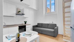 Un espacio habitable compacto con kitchenette, un sofá gris, una mesa de comedor con una botella y vasos, y un ventanal con vistas.