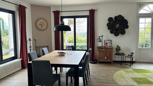 A dining area with a wooden table and chairs, a clock on the wall, and a view through the windows. - Le Clos des Pères House: 120 m², 3 bedrooms, 2 bathrooms, fenced-in yard. Pets allowed (Monflanquin)