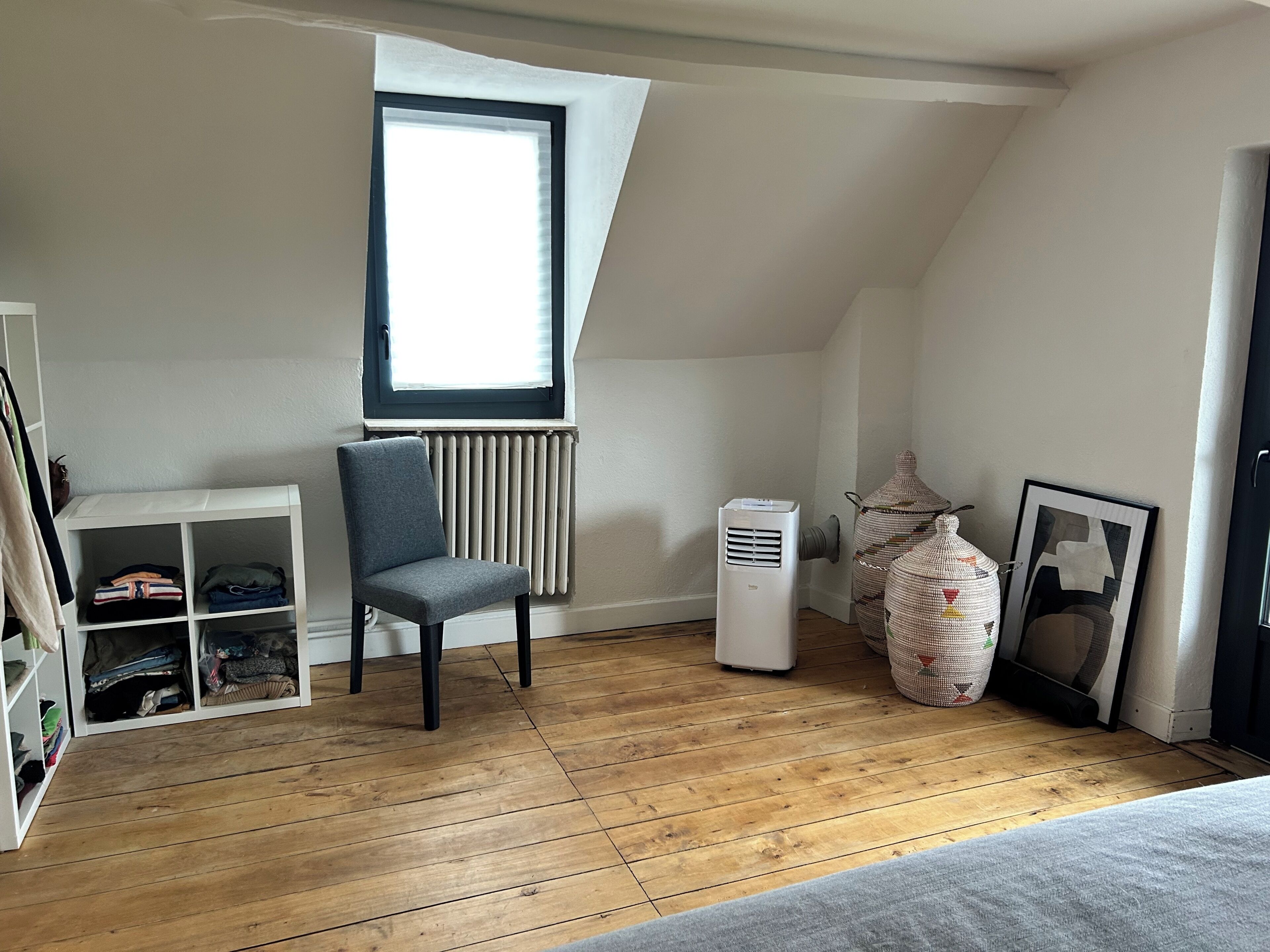 Een kamer met een radiator, een stoel, een ventilatie rooster, een plank met opgevouwen kleding en een raam.