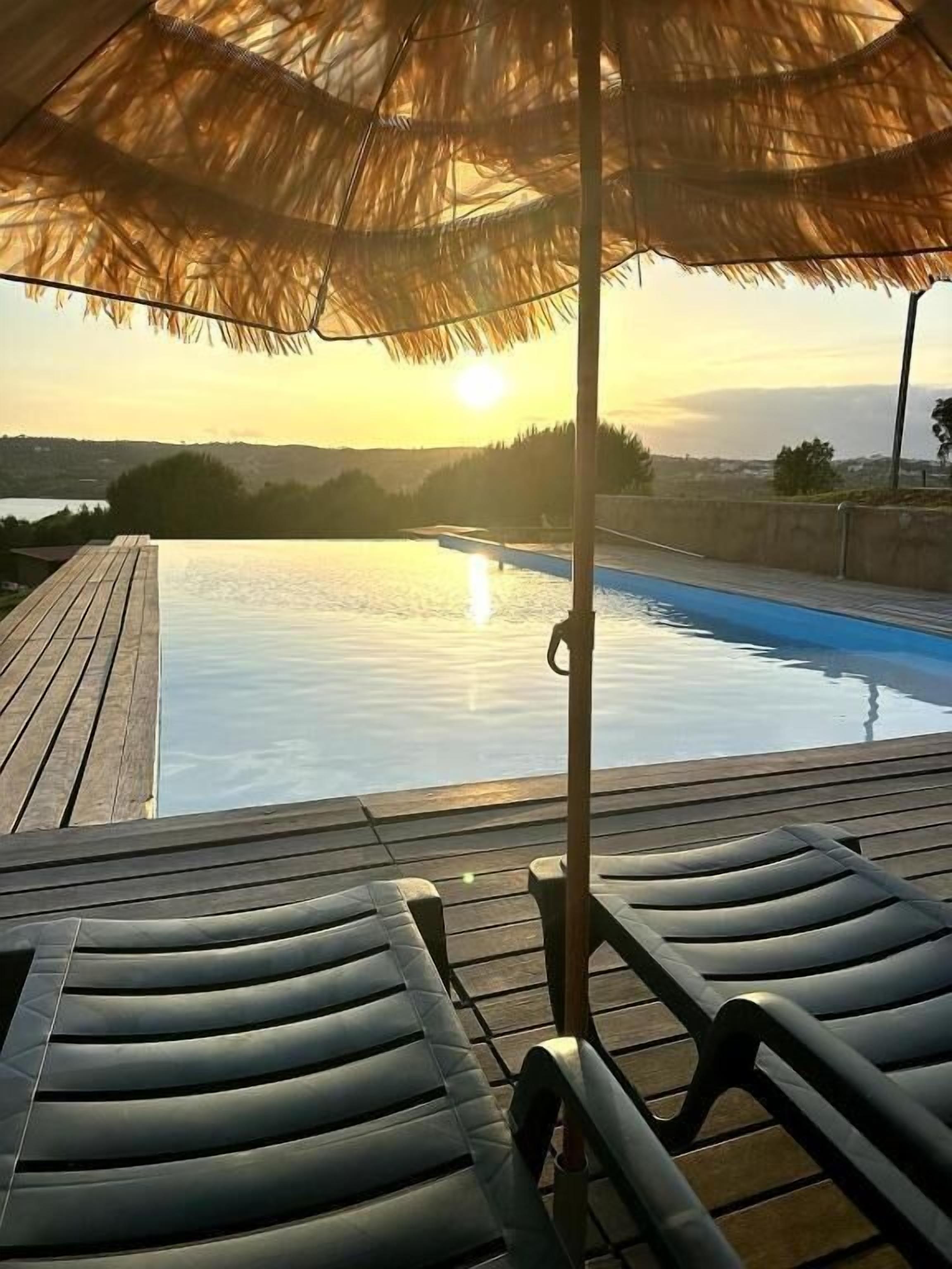 Una piscina infinita con terraza de madera, sillas de descanso, sombrilla de paja y vista al atardecer.