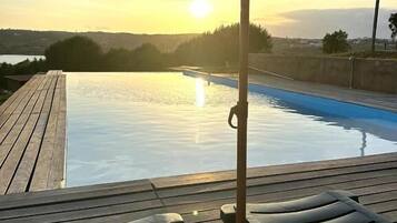 Una piscina infinita con terraza de madera, sillas de descanso, sombrilla de paja y vista al atardecer.