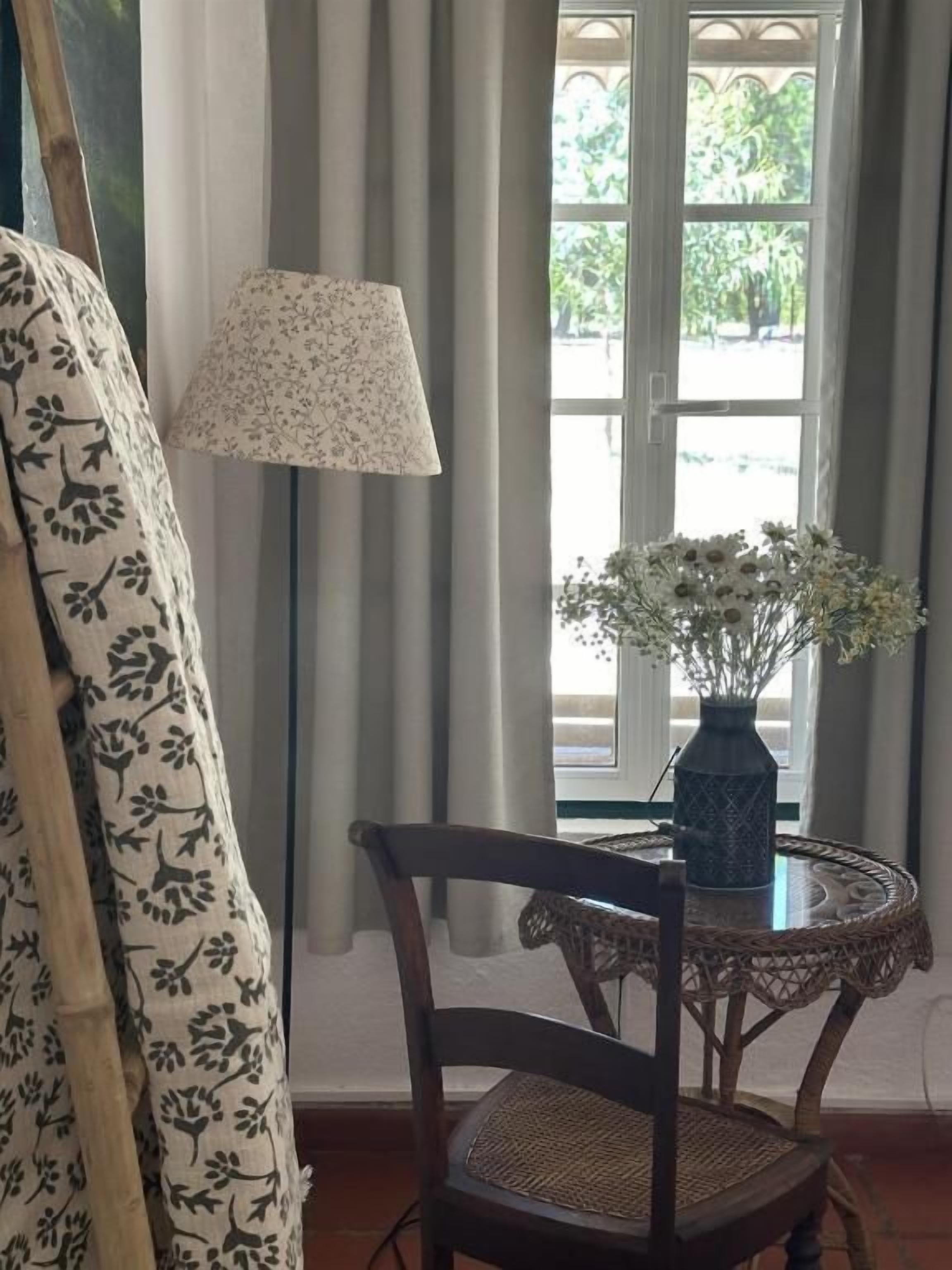 A room with a patterned curtain, a chair, a table with a vase of flowers, and a lamp.