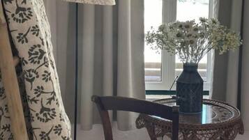 A room with a patterned curtain, a chair, a table with a vase of flowers, and a lamp.