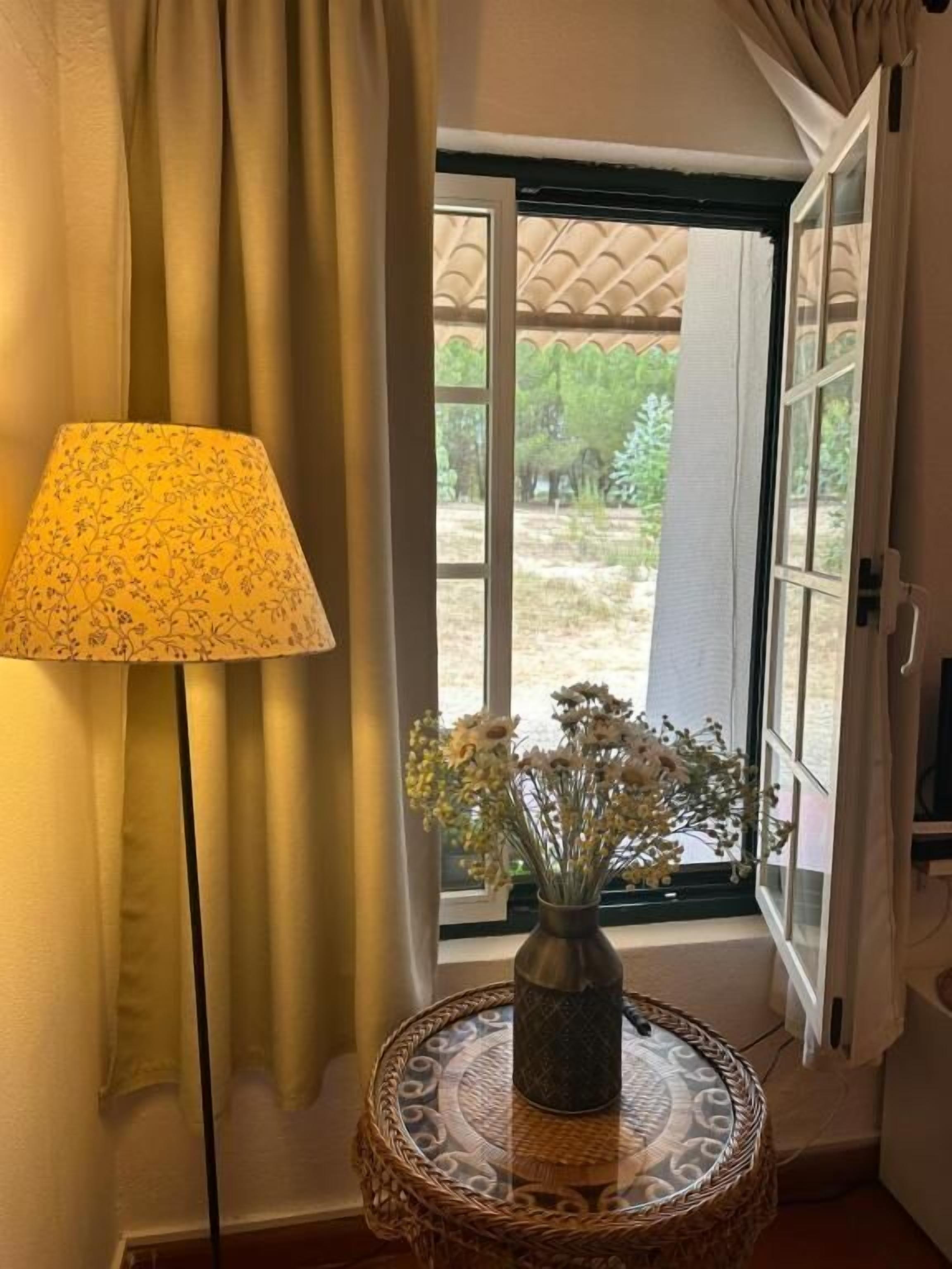 A room with a window, a lamp, and a vase with flowers on a round table.