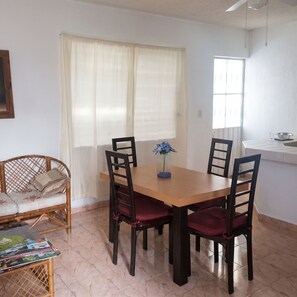 A dining area with a wooden table and four chairs, a wicker sofa, and a window with curtains.
