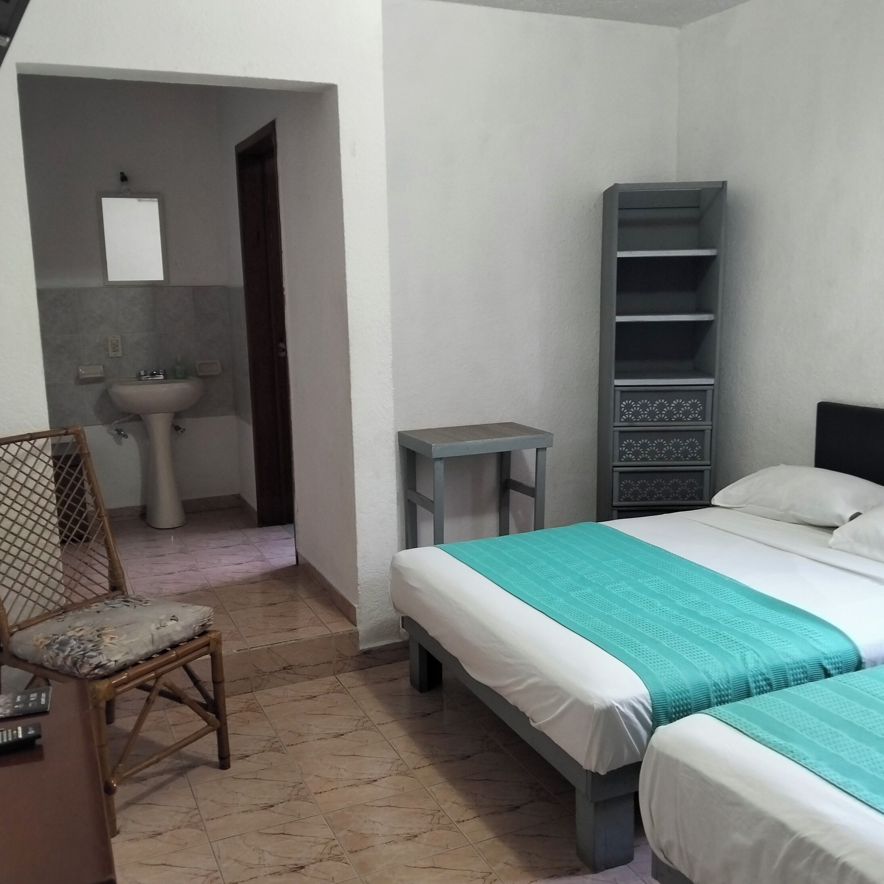 A bedroom with a bed, a chair, a bedside table, a shelf unit, and a bathroom with a sink.