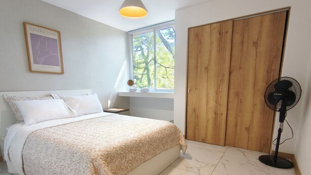 A bedroom with a bed, a wooden wardrobe, a standing fan, and a window with a view of trees. - Bright 3BR Oasis • 500Mbps WiFi & 24/7 Security (Medellín)