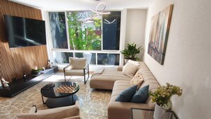 A modern living room with a sofa, chairs, a coffee table, a TV, and a large window with a view of greenery. - Bright 3BR Oasis • 500Mbps WiFi & 24/7 Security (Medellín)