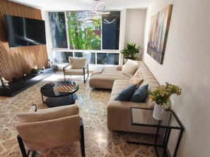 A modern living room with a sofa, chairs, a coffee table, a TV, and a large window with a view of greenery. - Bright 3BR Oasis • 500Mbps WiFi & 24/7 Security (Medellín)