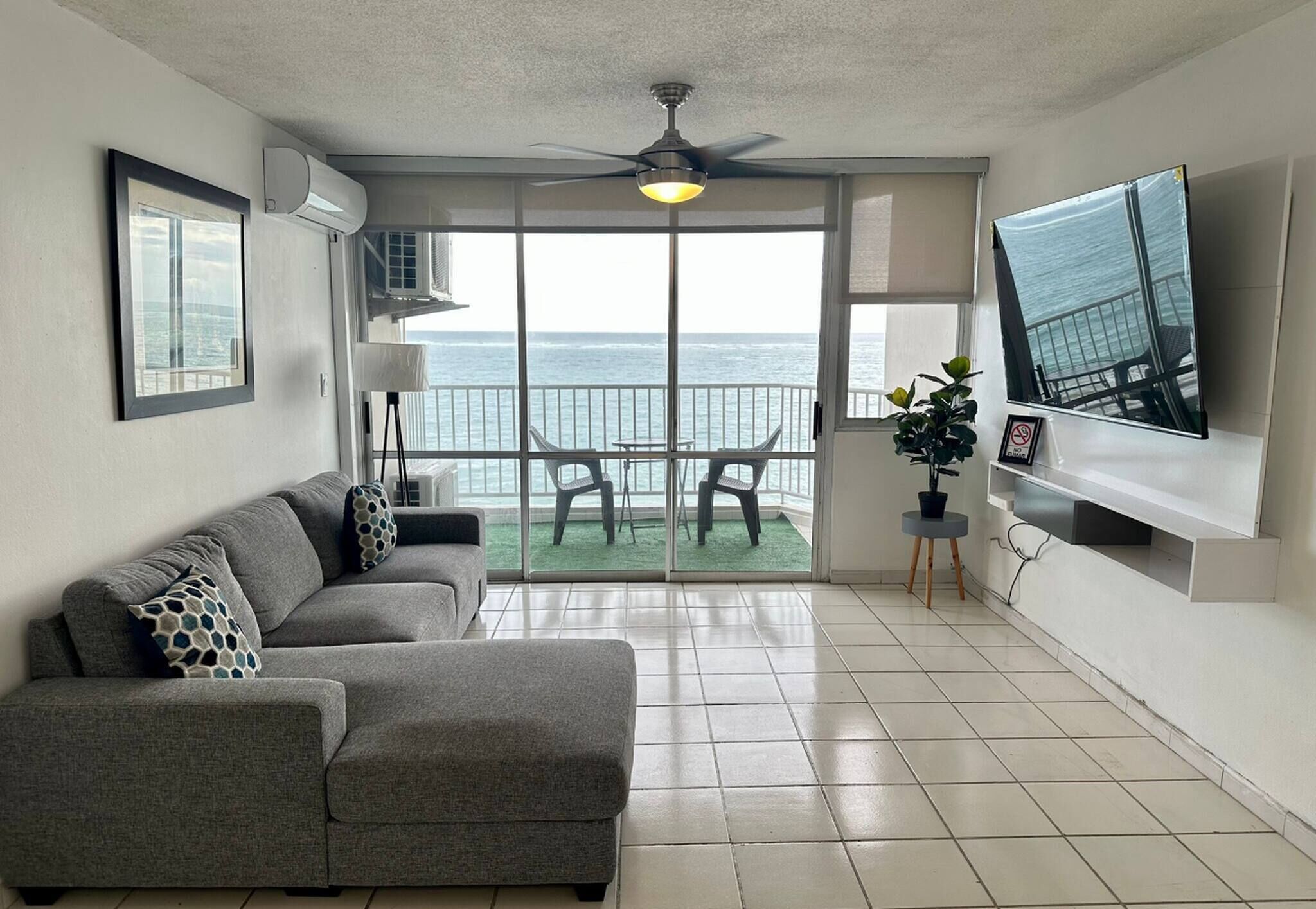 A living room with a gray sofa, a ceiling fan, a TV, and a balcony with a view of the ocean.