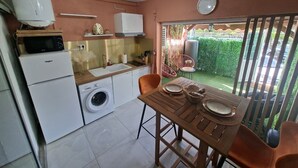 Kompakti keittiö, jossa on pesukone, mikroaaltouuni ja pesuallas. Keittiössä on ruokapöytä tuoleineen, ja lasiovesta avautuu näkymä puutarhaan.