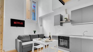 Eine moderne Küche mit Einbauofen, eine Essecke mit weißem Tisch und Stühlen, ein graues Sofa und ein an der Wand befestigter Fernseher mit der Aufschrift 'NETFLIX'.