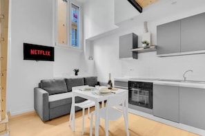 Eine moderne Küche mit Einbauofen, eine Essecke mit weißem Tisch und Stühlen, ein graues Sofa und ein an der Wand befestigter Fernseher mit der Aufschrift 'NETFLIX'.