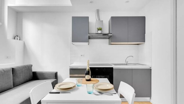 Eine moderne Küche mit grauen Schränken, einer weißen Arbeitsplatte und einem Essbereich mit weißem Tisch und grauem Sofa.