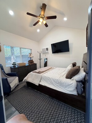 Ein Schlafzimmer mit Bett, Deckenventilator, Fernseher und einem Fenster mit Jalousien.